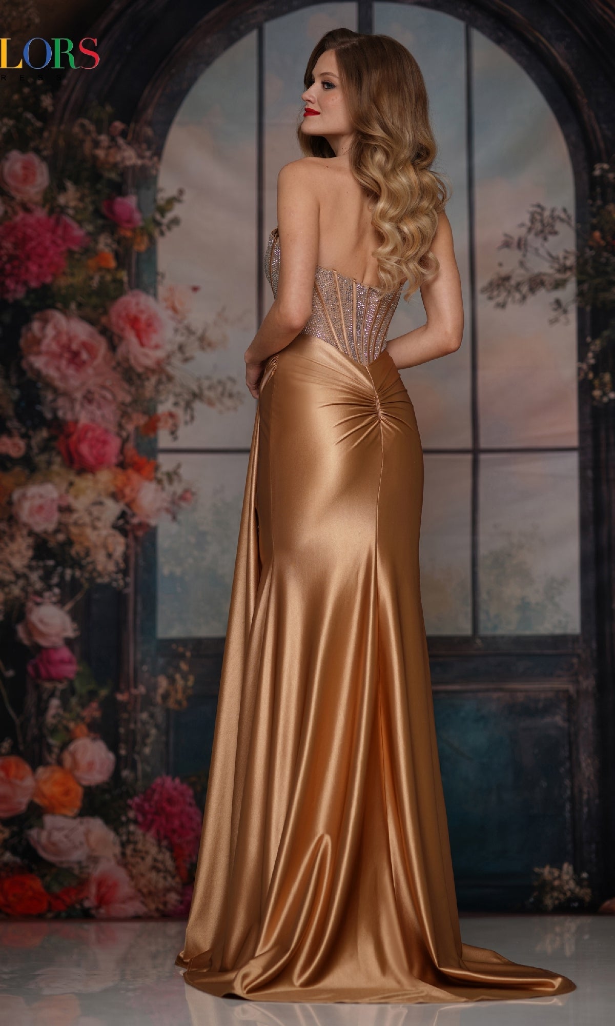 A woman with long wavy blonde hair wears the Strapless Long Corset Prom Gown 3933 by Colors Dress, posing gracefully before a floral background and arched window, smiling over her shoulder.